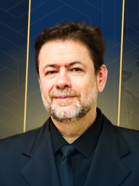 Fotografia de Edvaldo Luís Biancarelli, homem branco de cabelos castanho-escuros e barba grisalha aparada, com leve sorriso, vestindo terno, camisa e gravata pretos, diante de um fundo azul-marinho com linhas geométricas douradas.